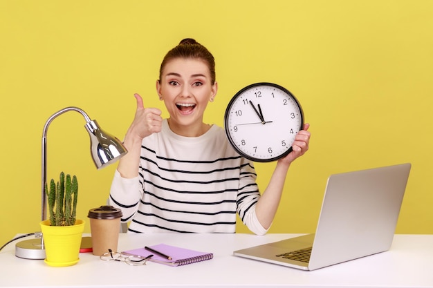 Travailleuse heureuse montrant les pouces vers le haut tenant une grande horloge murale regardant la caméra avec un sourire à pleines dents