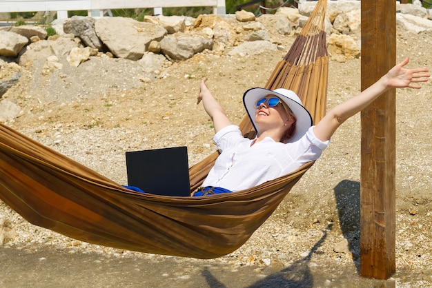 Travaillez de n'importe où jeune femme souriante pigiste en chapeau de paille travaillant sur ordinateur portable sur la plage