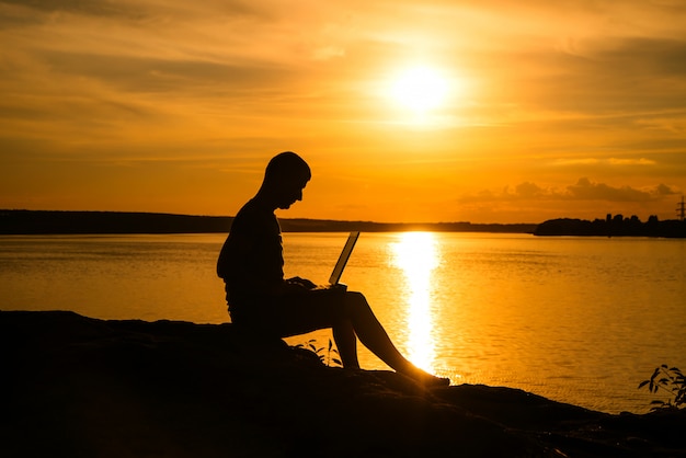 Travaillez en vacances avec un ordinateur portable près de la rivière au beau coucher de soleil.