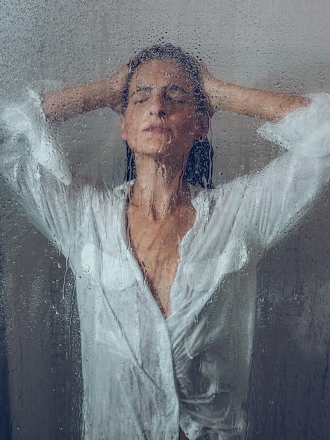 À travers le verre d'un charmant modèle féminin avec la main sur la tête se doucher dans une cabine de douche transparente avec porte mouillée pendant la routine quotidienne