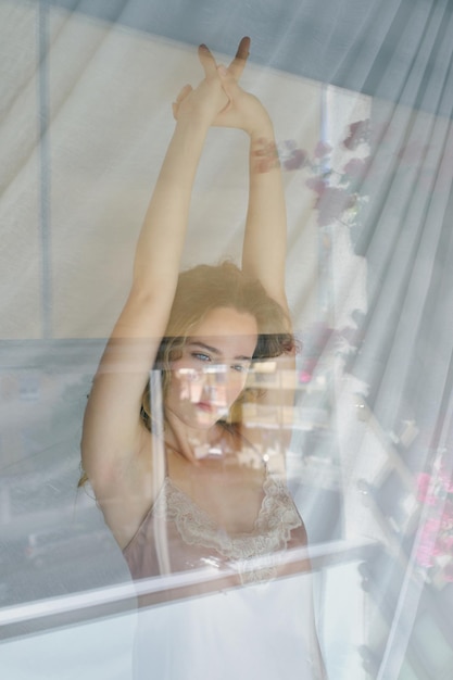 À travers la vue en verre d'une délicate jeune femme aux longs cheveux bouclés debout près de la fenêtre et levant les bras tout en s'étirant après s'être réveillée le matin