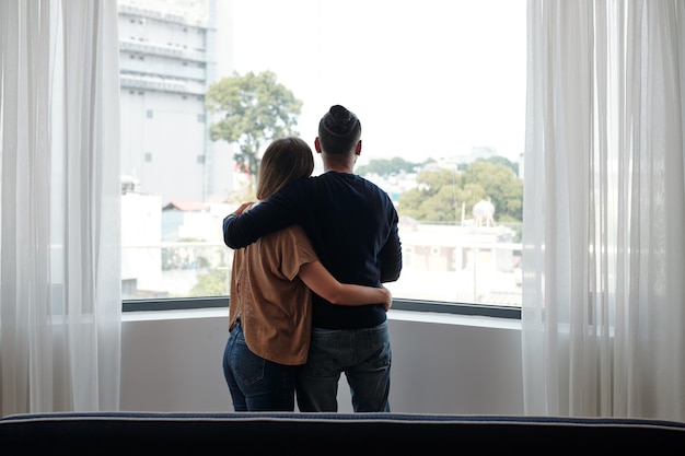 Étreignant un jeune couple debout devant une grande fenêtre et regardant à l'extérieur, vue de l'arrière