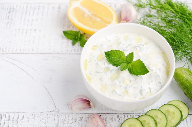 Trempette grecque ou vinaigrette tzatziki et ingrédients décorés à l’huile d’olive et à la menthe sur une table en bois blanche