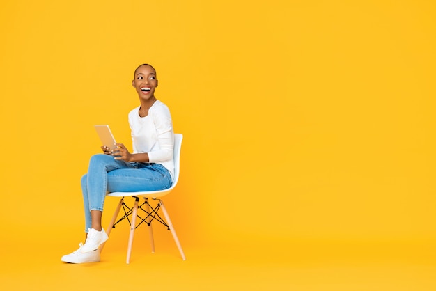 Photo trendy smiling african american woman sitting on a chair using tablet computer thinking et en regardant l'espace vide à côté du mur jaune isolé