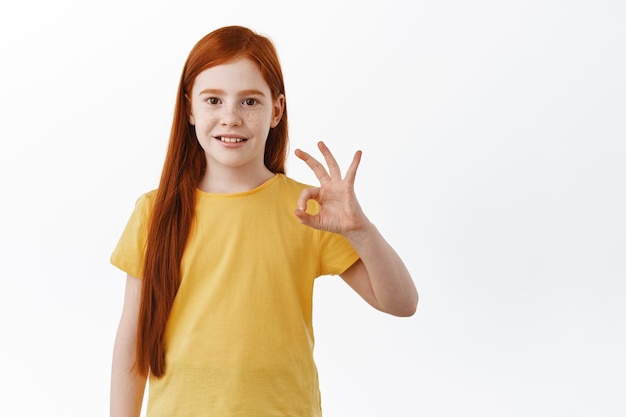 Très bien. Jolie petite fille aux longs cheveux roux et aux taches de rousseur montre un signe d'accord, sourit et dit oui, loue quelque chose d'excellent, une chose cool, hoche la tête en signe d'approbation, fond blanc