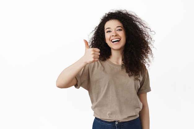 Très bien. Joyeuse fille brune souriante, hochant la tête en signe d'approbation, montrant les pouces vers le haut pour aimer et louer votre choix, complimenter l'excellent travail, recommander quelque chose, debout sur fond blanc