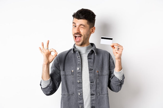 Très bien. Un mec souriant avec une carte de crédit en plastique montrant un signe correct, souriant et un clin d'œil satisfait, recommande la banque, debout sur fond blanc