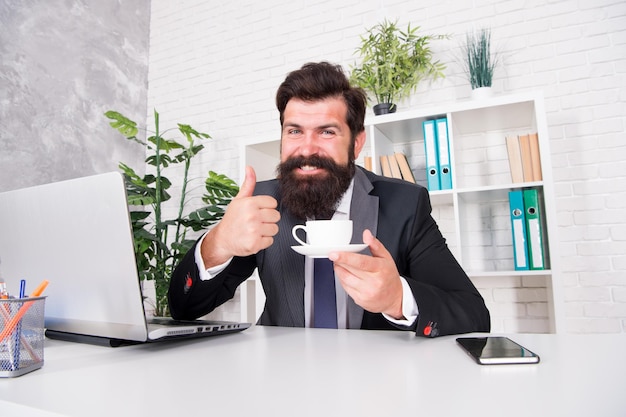 Très bon cappuccino Discuter des affaires autour d'un café Homme beau patron assis au bureau en buvant du café Espace de travail confortable Homme d'affaires barbu costume formel relaxant avec du café Routines de la vie de bureau