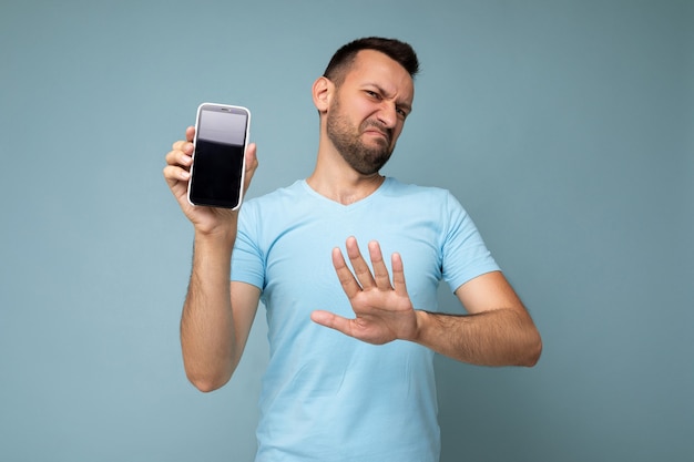 Triste beau jeune homme brunet non rasé portant un t-shirt bleu de tous les jours isolé sur fond bleu