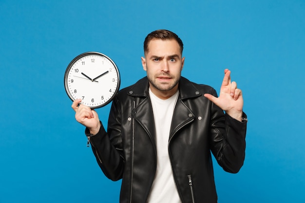 Triste bouleversé élégant jeune homme mal rasé en t-shirt blanc veste en cuir noir tenant une horloge ronde isolée sur fond de mur bleu portrait en studio. Concept de mode de vie des gens. Dépêche-toi. Maquette de l'espace de copie.