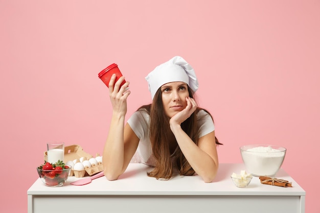 Triste chef cuisinier confiseur ou boulanger en t-shirt blanc tablier, chapeau de chefs toque assis à table avec une tasse de café en papier isolé sur fond rose pastel en studio. Maquette du concept de nourriture de l'espace de copie.