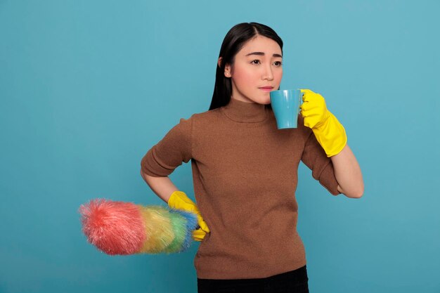 Triste contrarié et déprimé d'une jeune femme de ménage prenant du café dans le gant jaune avec une brosse à poussière colorée d'autre part isolée sur fond bleu, Nettoyage de la maison, Temps de pause-thé de rafraîchissement