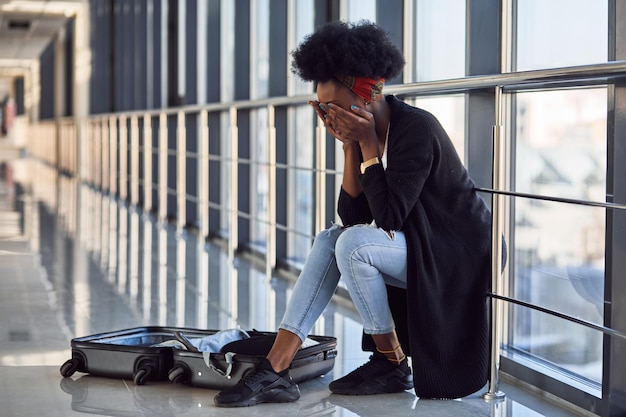 Triste et déprimée jeune passagère afro-américaine en tenue décontractée assise à l'aéroport avec des bagages.