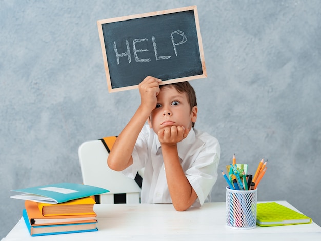 Triste écolier frustré et fatigué à la table et tenant du papier avec word Help. Difficultés d'apprentissage, concept d'éducation