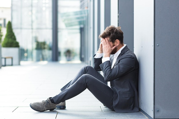 Triste entrepreneur déprimé en costume formel homme travailleur assis près de la rue en plein air près du centre d'affaires de bureau moderne. Un homme d'affaires bouleversé a perdu son emploi en raison d'une crise financière. Un employé a un problème. à l'extérieur