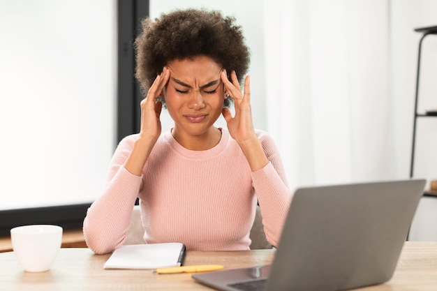 Triste femme afro-américaine millénaire fatiguée avec un ordinateur portable souffrant de maux de tête presse les mains vers les tempes