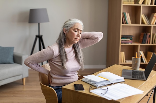 Triste femme âgée aux cheveux gris européenne fatiguée faisant un massage du dos travaillant avec un ordinateur portable dans le salon