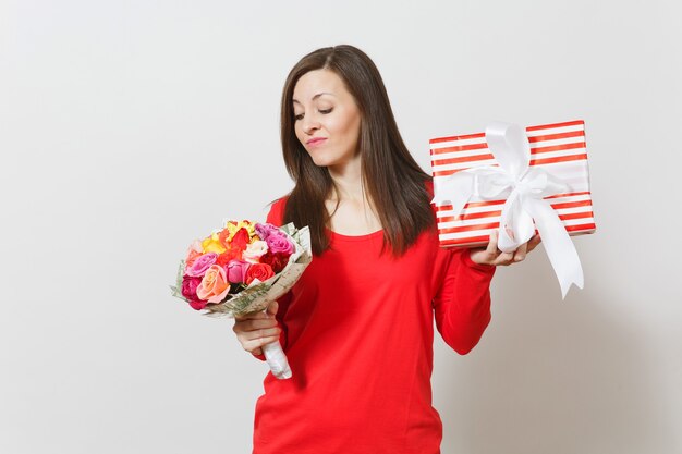 Triste femme bouleversée en vêtements rouges tenant un bouquet de belles fleurs roses, boîte présente avec cadeau isolé sur fond blanc. Saint-Valentin, concept de vacances d'anniversaire de la Journée internationale de la femme.