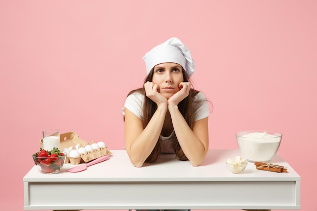 Triste femme chef cuisinier confiseur ou boulanger en t-shirt blanc tablier, chapeau de chefs toque cuisson gâteau ou cupcake à table isolé sur fond pastel rose en studio. Maquette du concept de nourriture de l'espace de copie.