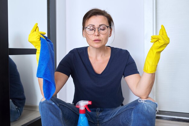 Triste femme méditant fatiguée dans des gants avec un détergent et un chiffon assis sur le sol dans la chambre a fini de nettoyer la maison. Hygiène, pureté, maison, concept de propreté