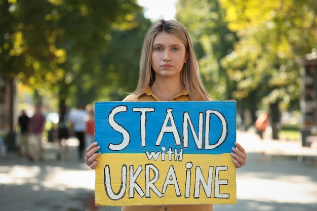 Triste femme tenant une affiche aux couleurs du drapeau national et des mots Stand avec l'Ukraine à l'extérieur