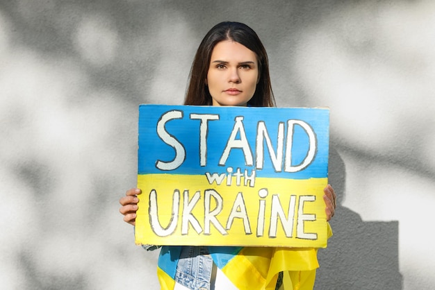 Triste femme tenant une affiche aux couleurs du drapeau national et des mots Stand avec l'Ukraine près du mur lumineux