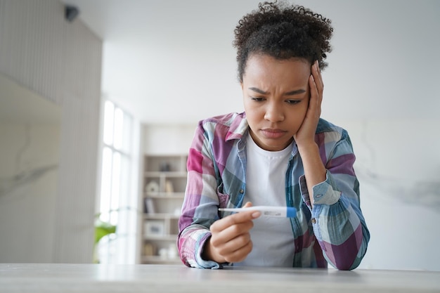 Triste fille afro-américaine regarde le thermomètre a un symptôme covid de fièvre à température corporelle élevée