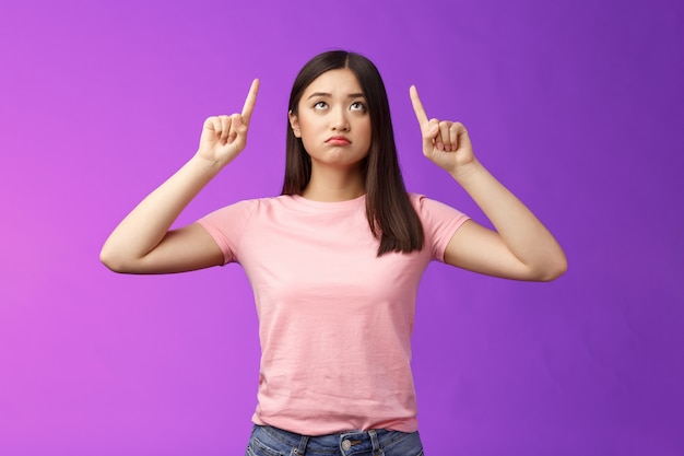 Triste fille asiatique boudante déçue avec une courte coupe de cheveux sombre, l'air pointant vers le haut, contrariée, jalouse, regrettant, ratant une bonne opportunité, regardant la publicité supérieure malheureuse, debout sur fond violet.