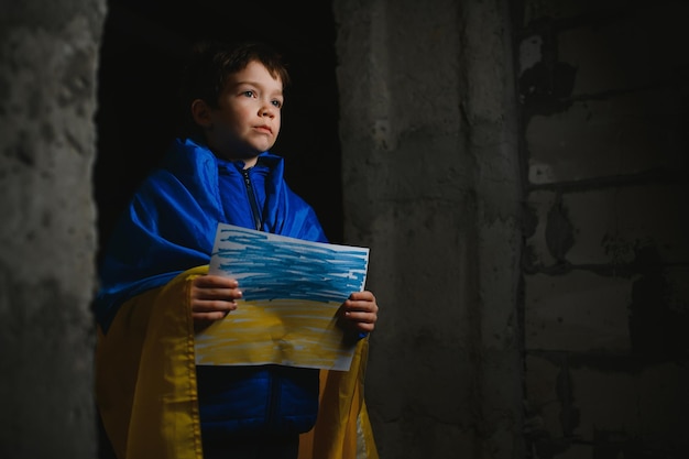 Un triste garçon ukrainien enveloppé dans un drapeau ukrainien se cachant des bombardements dans un abri anti-bombes dans la ville de Marioupol ou de Kiev L'agression de la Russie La guerre en Ukraine et 2022