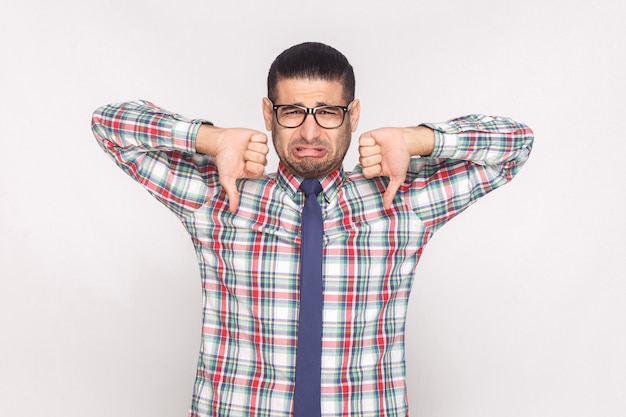 Triste homme d'affaires barbu malheureux en chemise à carreaux colorée, cravate bleue et lunettes debout et regardant avec le pouce levé et n'aimant pas le geste. tourné en studio intérieur, isolé sur fond gris clair.