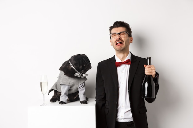 Triste homme pleurant et buvant du champagne dans une bouteille tout en carlin en costume de fête mignon regardant confus, debout sur fond blanc.