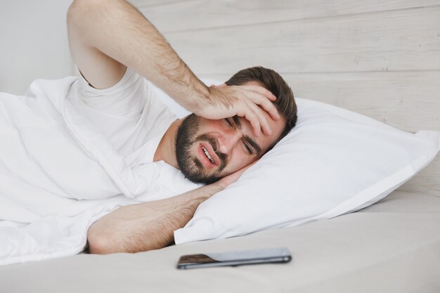 Triste jeune bel homme barbu allongé dans son lit avec une couverture d'oreiller en drap blanc dans la chambre à la maison