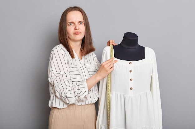 Triste jeune femme adulte insatisfaite debout près d'un mannequin avec une nouvelle robe isolée sur fond gris regardant la caméra avec une expression confuse tenant un ruban à mesurer