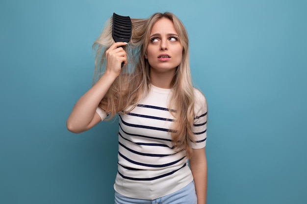 Triste jeune femme blonde bouleversée avec difficulté à se peigner les cheveux qui tombent sur un fond bleu