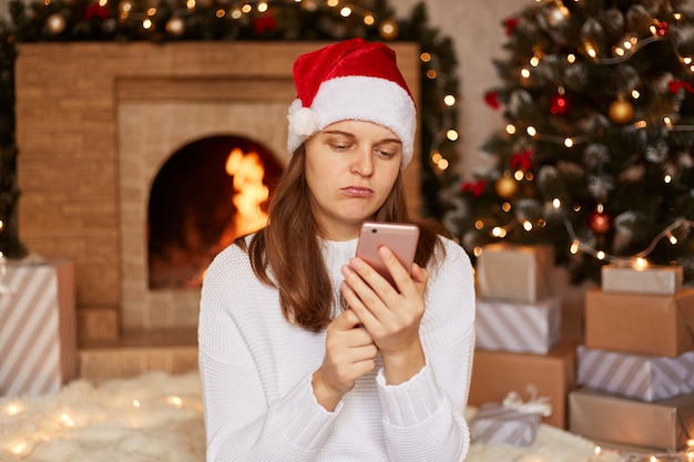 Triste jeune femme caucasienne aux cheveux noirs portant un bonnet de Noel et un pull utilisant un smartphone avec une expression faciale contrariée, assise dans un salon festif avec cheminée et arbre de Noël et tapant un message.