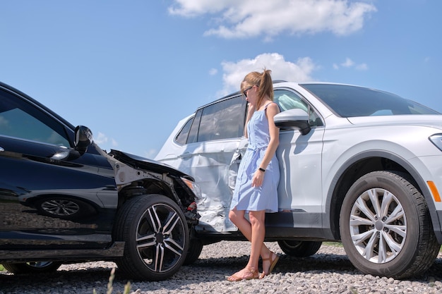 Triste jeune femme conductrice debout près de sa voiture brisée, l'air choquée par des véhicules accidentés dans un accident de la route