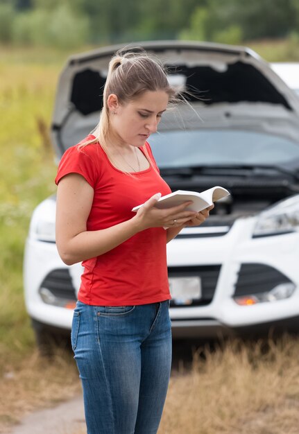 Triste jeune femme lisant le manuel du propriétaire à la voiture cassée dans le pré