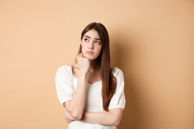 Triste jeune femme à la recherche pensive dans le coin supérieur gauche, se sentant bouleversée ou en détresse, debout seule sur fond beige.