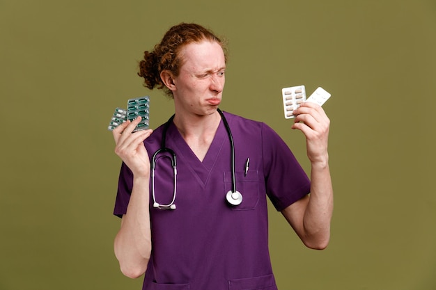 Triste jeune médecin de sexe masculin en uniforme avec stéthoscope tenant et regardant des pilules isolées sur fond vert