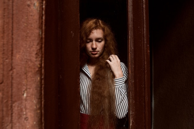 Triste modèle aux cheveux rouges debout derrière une porte vitrée fermée. Gouttes de pluie sur le verre