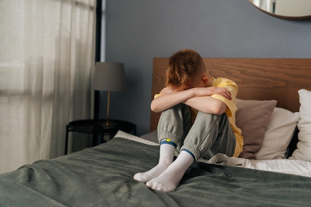 Triste petit garçon méconnaissable étreignant les genoux sanglotant la tête baissée et pleurant assis sur le lit dans la chambre des enfants Enfant stressé ressentant de la pression