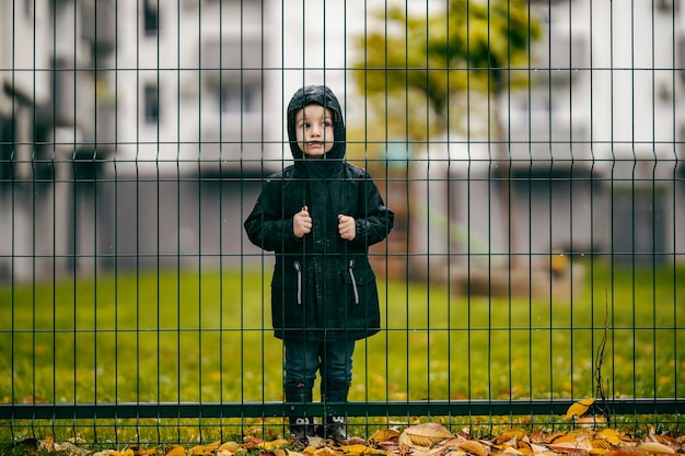 Un triste petit par se tient sur la pluie à côté d'une clôture sur la pluie et regarde entre les grilles