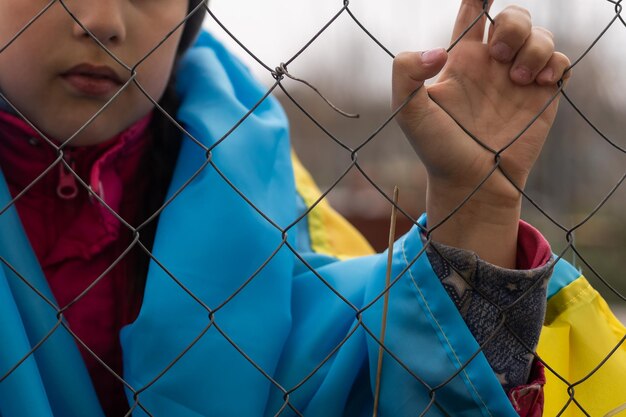 Triste petite fille avec le drapeau de l'Ukraine derrière une clôture métallique. Problème social des réfugiés et des migrants forcés