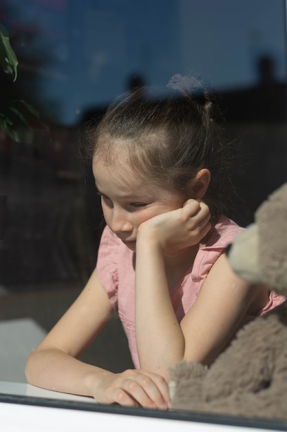 Triste petite fille avec son ours en peluche à la fenêtre.