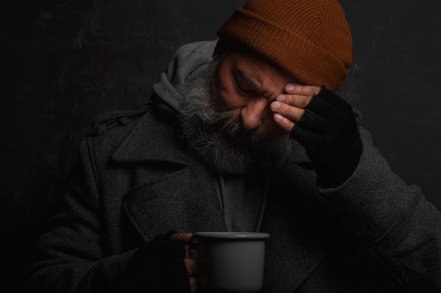 Triste sans-abri avec une barbe grise tenant une tasse de thé chaud pour se réchauffer par une nuit froide