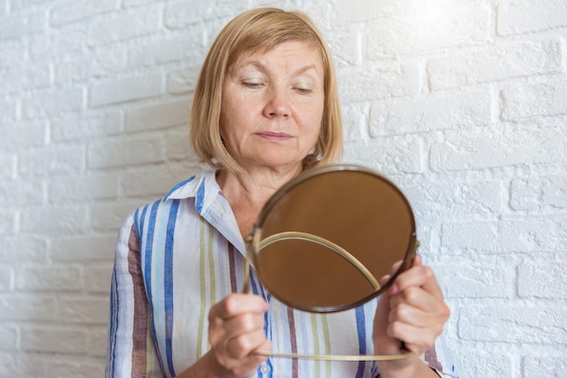 Triste vieille femme mature senior regardant la réflexion du visage dans un miroir rond. Vie domestique, spa à domicile, soins personnels et concept de soins