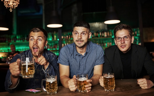 Trois amateurs de sport dans un bar regardent le football. Avec de la bière dans les mains.