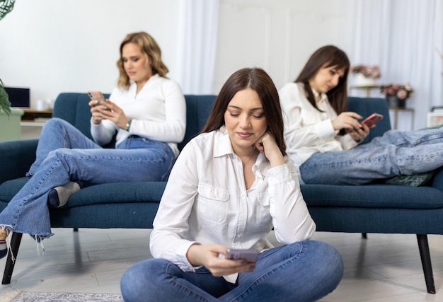Trois amies en chemises et jeans s'assoient sur et près du canapé dans la chambre et grimpent sur les réseaux sociaux
