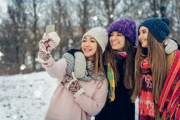 Trois amies à l'extérieur dans des chapeaux tricotés à l'aide d'un téléphone portable par temps froid et neigeux