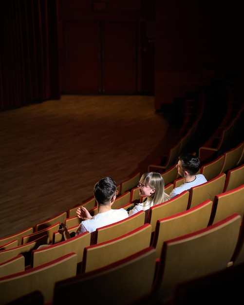 Trois amis, une adolescente et deux garçons souriants regardent un film dans un cinéma vide avec du rouge